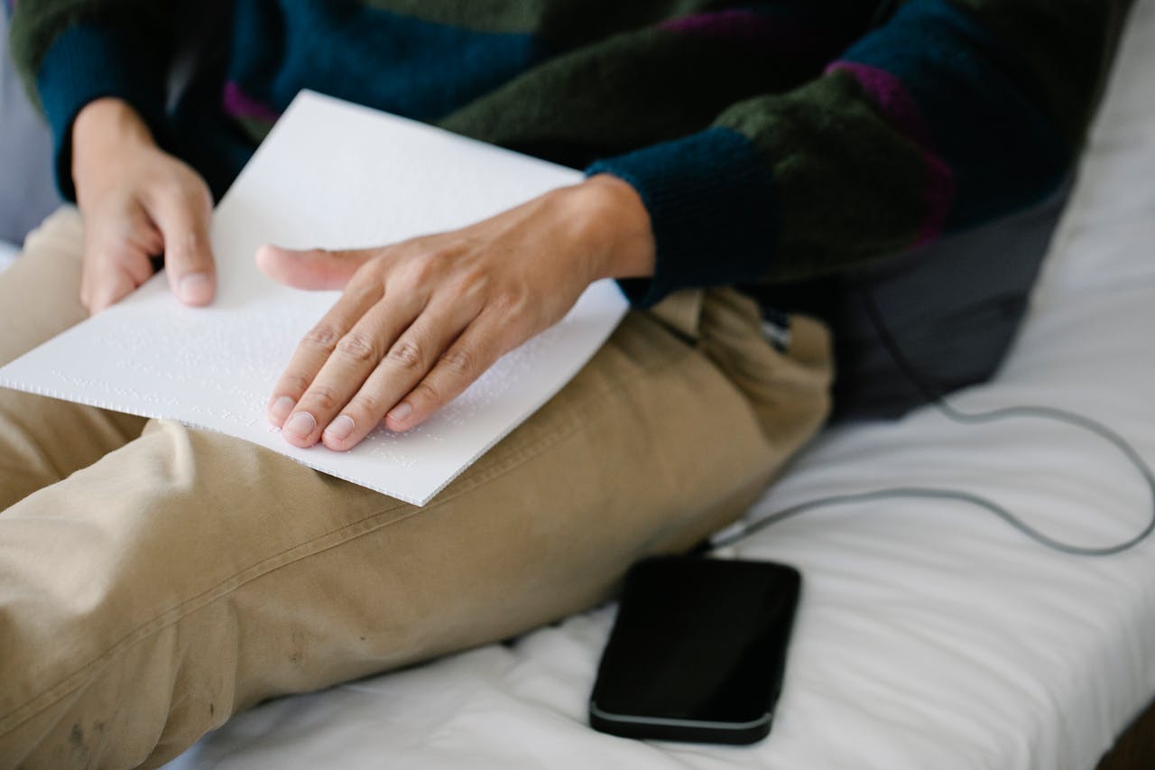 about-us A person reads Braille text while sitting on a bed with a smartphone nearby.